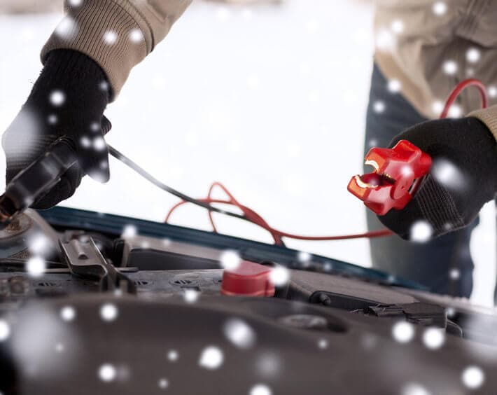 Two gloved hands attaching jumper cables onto dead car battery, while snow falls gently