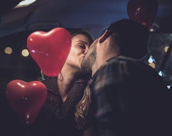 Couple kissing in vehicle with red heart shaped balloons around them.