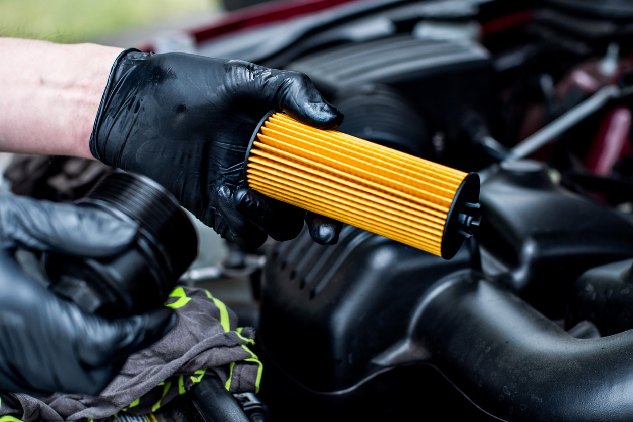 Image of person wearing black gloves holding oil filter with under hood of vehicle in backdrop